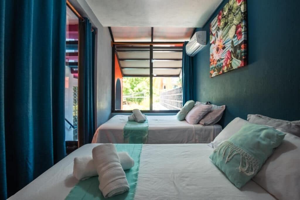 Habitación de hotel con dos camas, una ventana con cortinas, aire acondicionado en la pared y un cuadro de colores.