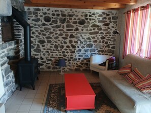Un mur de pierre, un canapé, un pouf rouge, une lampe et une fenêtre avec des rideaux.
