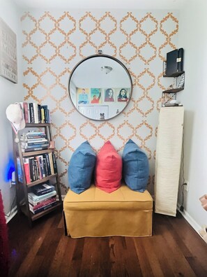 A room with a patterned wallpaper, a round mirror, a bookshelf, and a small bench with cushions.