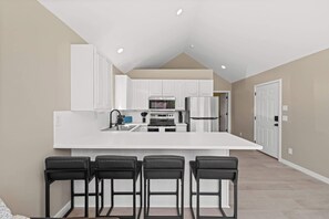 A modern kitchen with white cabinetry, a central island with bar stools, and recessed ceiling lighting. - Modern 1BR Retreat with Garage and Yard (Coeur d'Alene)