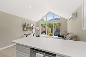 A modern kitchen with a large island, a dining area with a table and chairs, and a view of a residential street through large windows. - Modern 1BR Retreat with Garage and Yard (Coeur d'Alene)