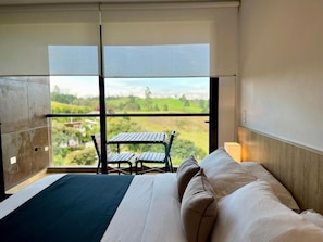 A hotel room with a bed, a view of a green landscape, and a small table and chair outside the window.