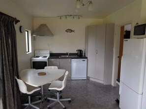 A compact kitchen with a stove, sink, and refrigerator. There is a dining table with chairs. - Hosts on the Coast Our Little Gem (Whitianga)