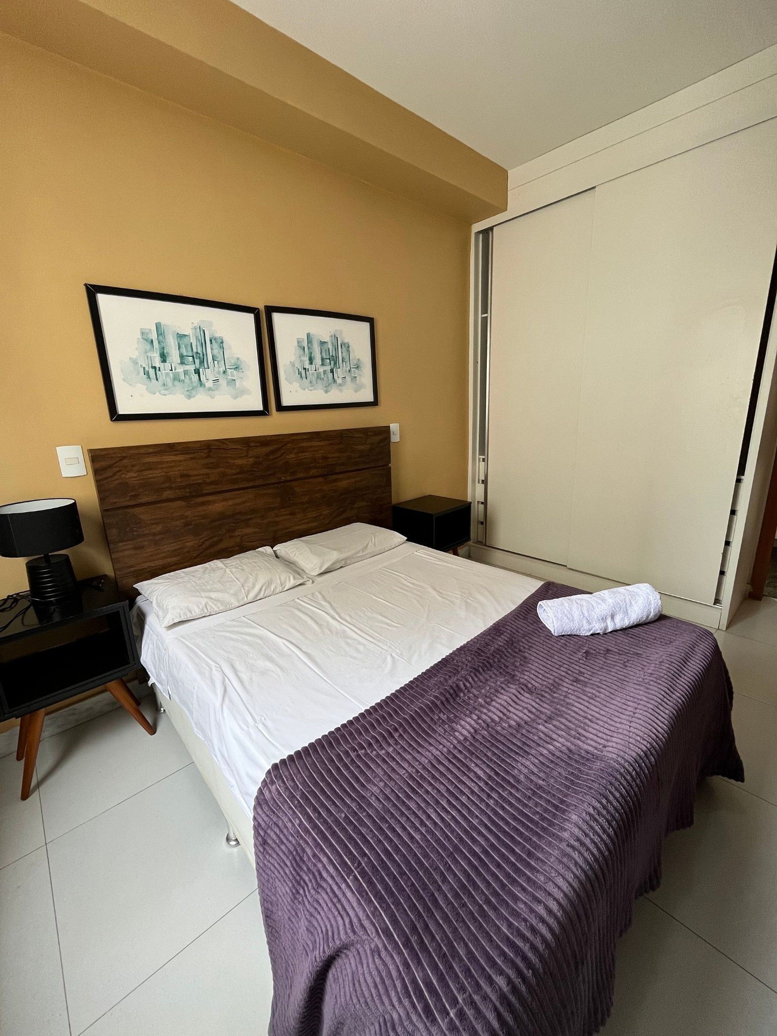 Una habitación de hotel con una cama, una mesita de noche, una lámpara y dos cuadros con vistas urbanas en la pared.