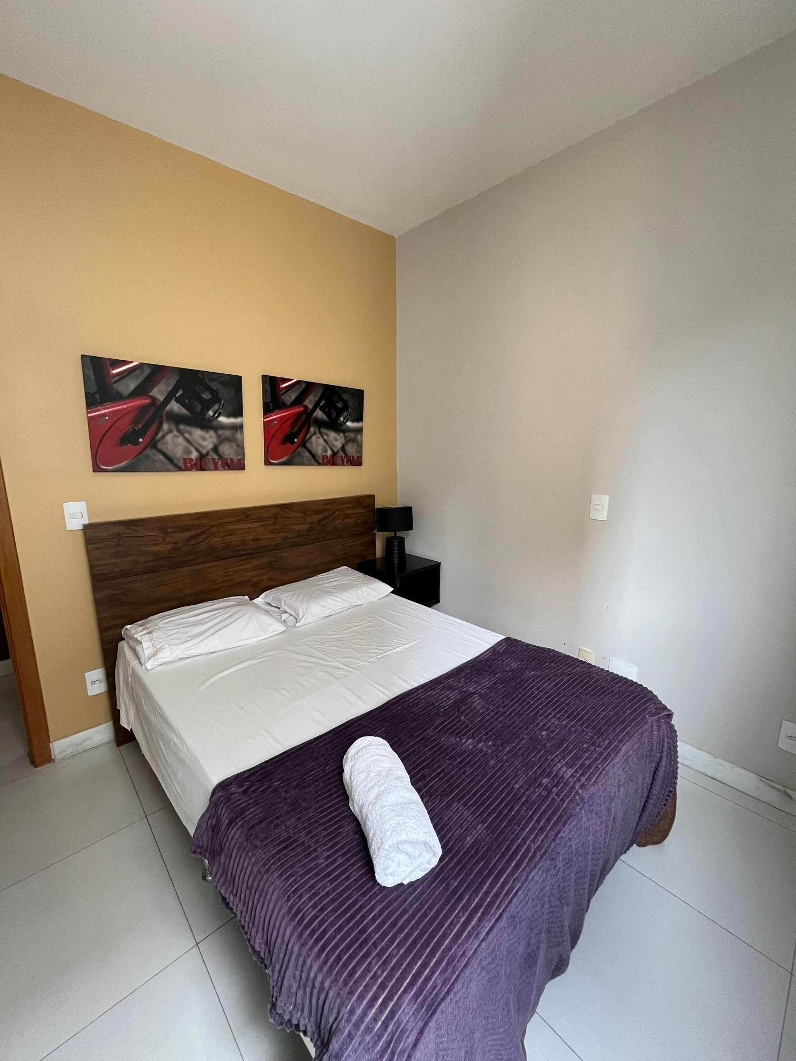 A neatly made bed with a purple blanket and a rolled towel, two wall-mounted artworks depicting bicycles, and a wooden headboard.