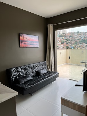 Un salon moderne avec un canapé en cuir noir, un tableau, une table en verre et un balcon offrant une vue sur la ville.