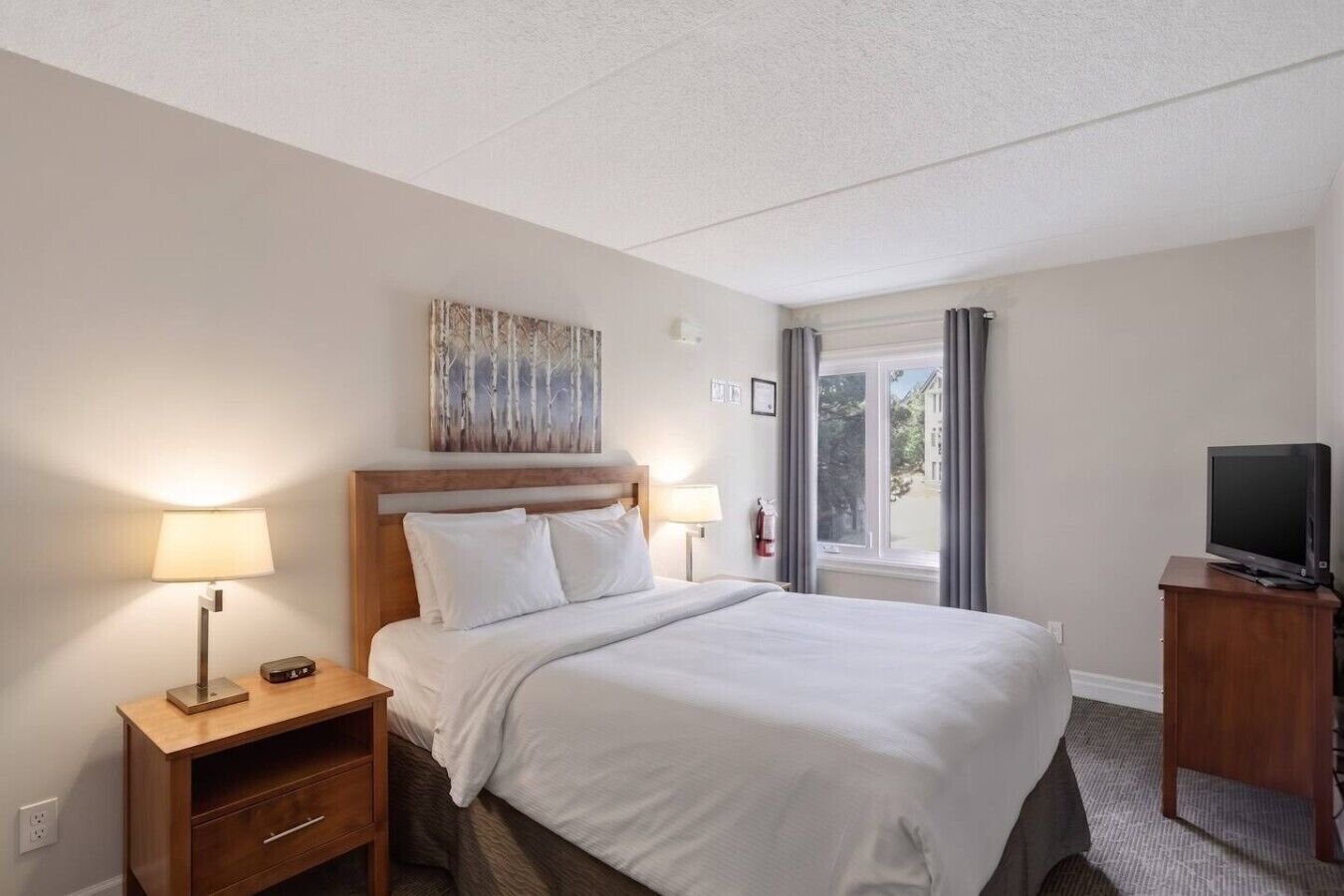 A hotel room with a bed, bedside lamps, a television on a stand, a painting above the bed, and a window with curtains.