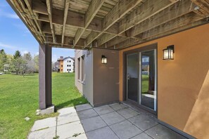 Ein überdachter Außenbereich mit Betonboden, Holzboden und einer Glastür, die zu einem Gebäude mit brauner Fassade führt.