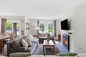A living room with a fireplace, a television mounted on the wall, and a dining area with a table and chairs. - Village Side 3BDR | New Kitchen & Private Patio (The Blue Mountains)