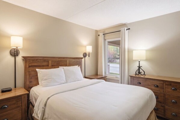 A neatly made bed with a wooden headboard, two bedside tables with lamps, a window with curtains, and a wooden dresser with drawers.