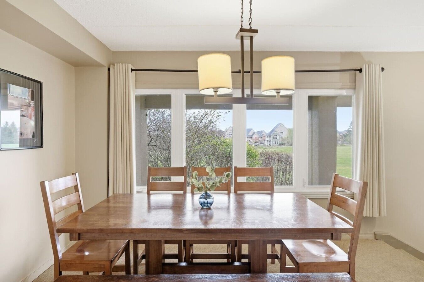 Une salle à manger avec une table en bois, quatre chaises, un vase rempli de fleurs et une vue sur les maisons, visible à travers de grandes fenêtres.