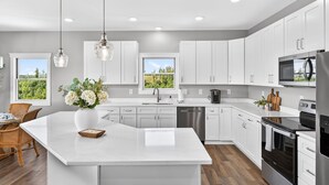 Una cocina moderna con armarios blancos, una isla grande, electrodomésticos de acero inoxidable y vistas al exterior.