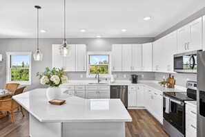 Une cuisine moderne avec des meubles blancs, un grand îlot central, des appareils en acier inoxydable et une vue sur la verdure environnante.