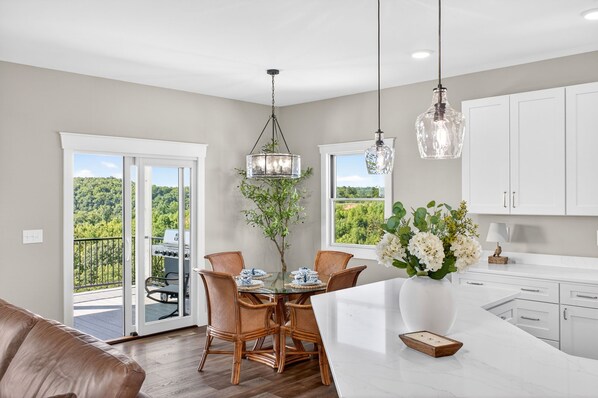 Une cuisine moderne avec des meubles blancs, un coin repas avec des chaises en osier, une table à plateau en verre et une vue sur la verdure grâce aux portes-fenêtres coulissantes.