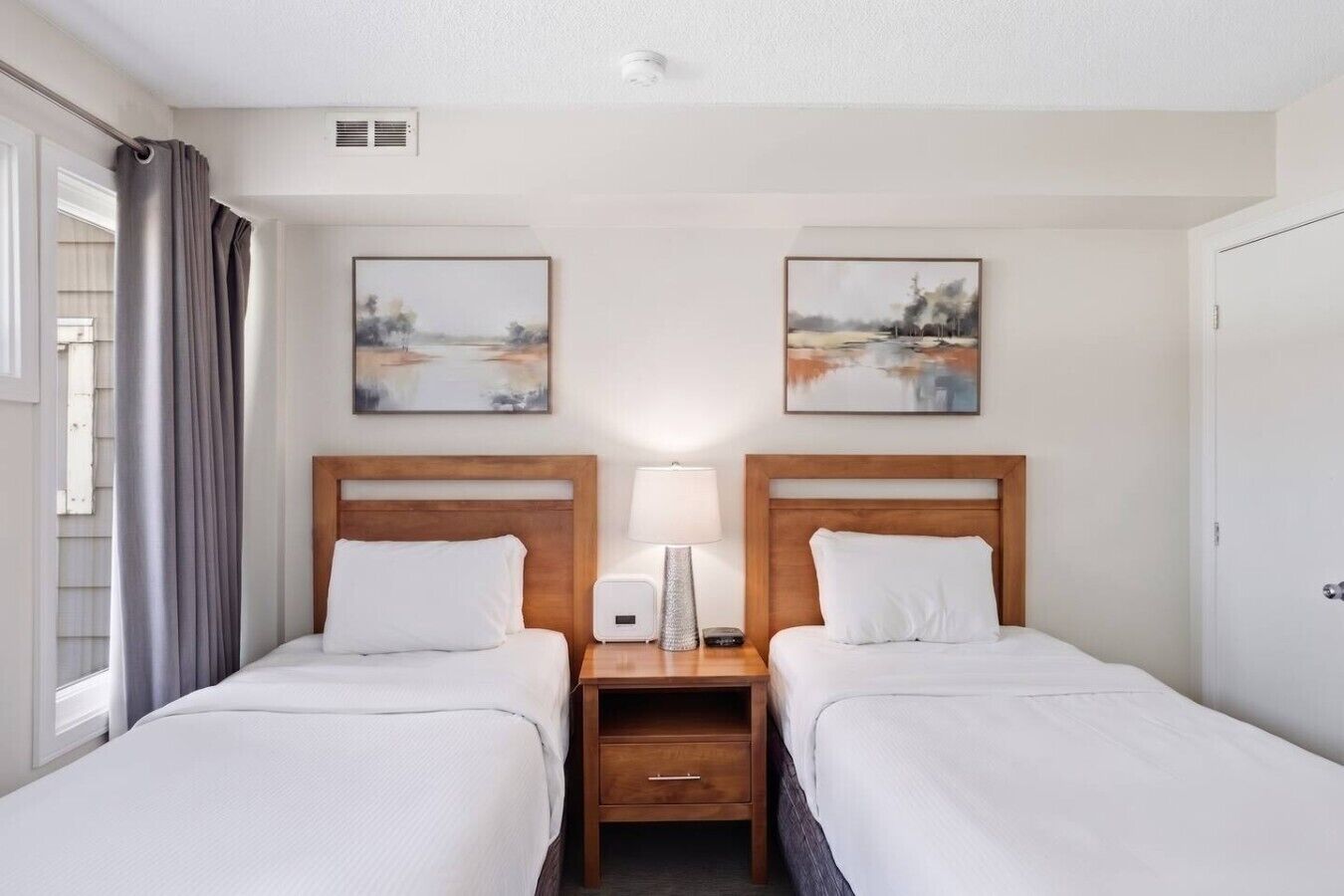 A hotel room with two beds, a nightstand with a lamp, and two paintings on the wall.