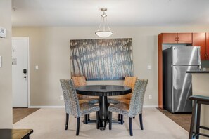 A dining area with a round table, four chairs, a painting on the wall, a ceiling light, and a stainless steel refrigerator.