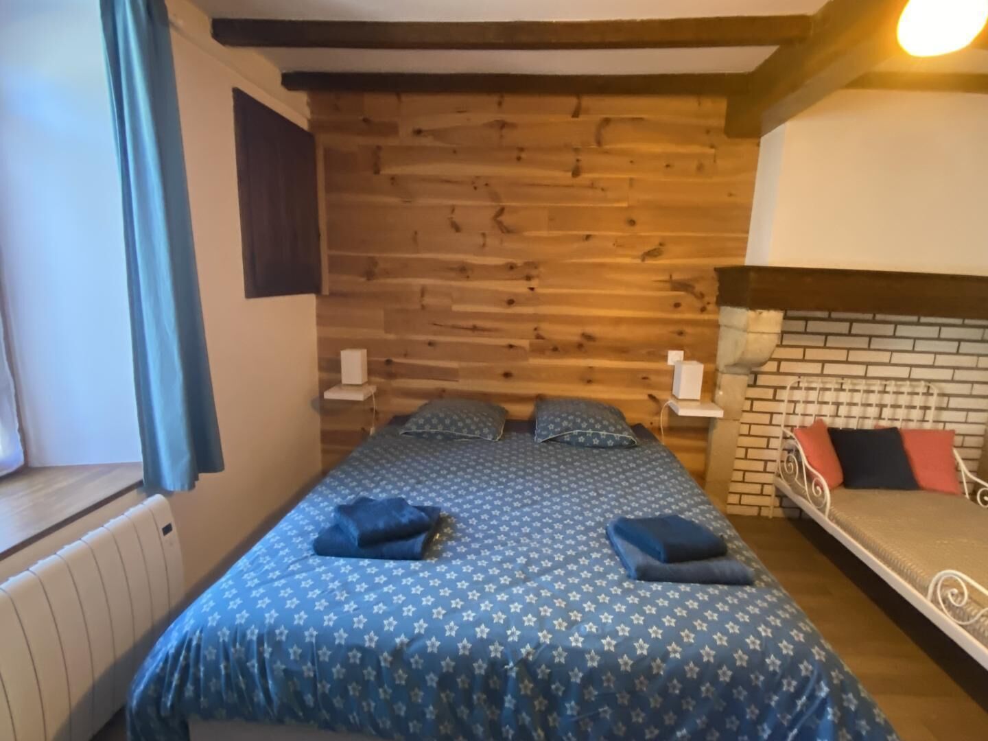 A room with a wooden wall, a bed with a patterned bedspread, a window with curtains, and a small table with a lamp.