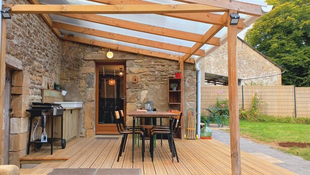 Un patio cubierto con una mesa y sillas de madera, un muro de piedra y una pequeña área de cocina con una parrilla.