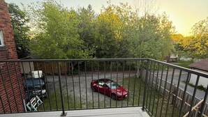 En altan med gelænder, en rød bil parkeret i baggrunden, og en murstensbygning til venstre.