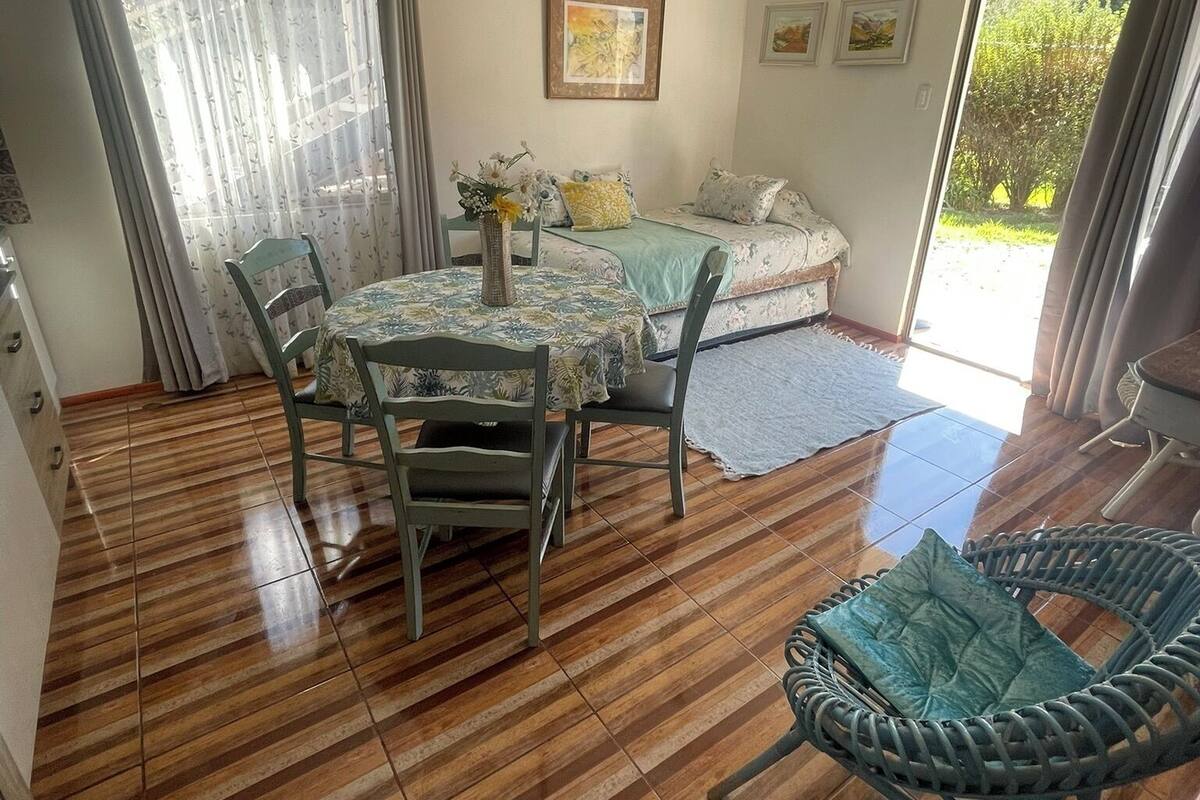 A room with a dining table, chairs, a sofa, a wicker chair, a ceiling fan, and a view of greenery outside.