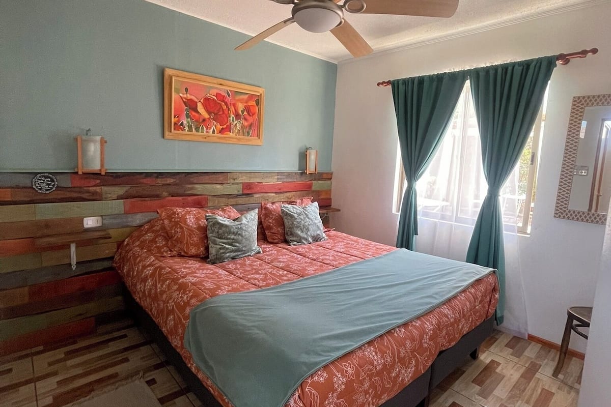 A bedroom with a bed, a ceiling fan, a window with curtains, a painting on the wall, and a wooden headboard.