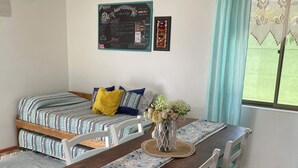 Una habitación con una mesa de comedor de madera, sillas blancas, un sofá con cojines estampados, un jarrón decorativo y una ventana con cortina.