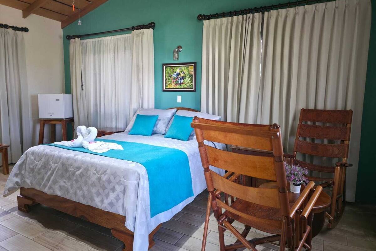 Un dormitorio con una cama, una silla de madera, una mesita con una planta en maceta y un televisor sobre un mueble.