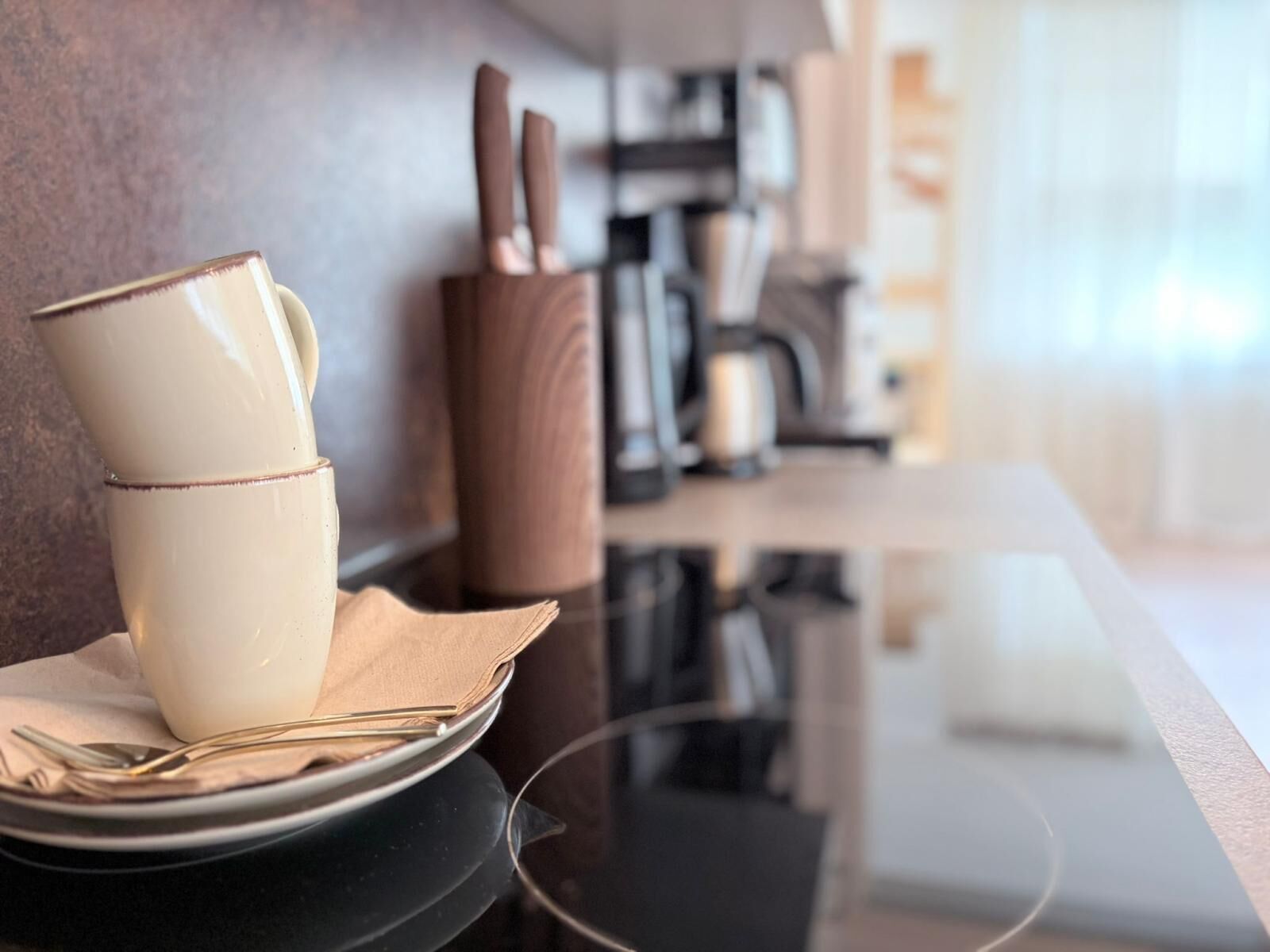 A modern kitchen with a coffee machine, knife block, and a stack of white mugs on a black countertop.