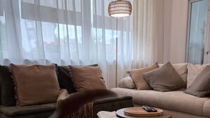 Una sala de estar con dos sofás, una mesa de centro redonda de madera, una lámpara de pie y un gran ventanal con cortinas translúcidas.