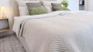 Una cama bien hecha con colcha, una lámpara de noche y una planta pequeña en la mesita.
