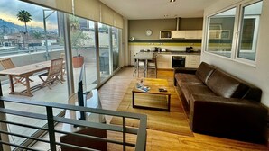 A modern apartment with a balcony, a living room with a sofa and coffee table, a kitchen with a microwave and stove, and large windows offering a city view. - Beautiful superior duplex apartment (Santiago)