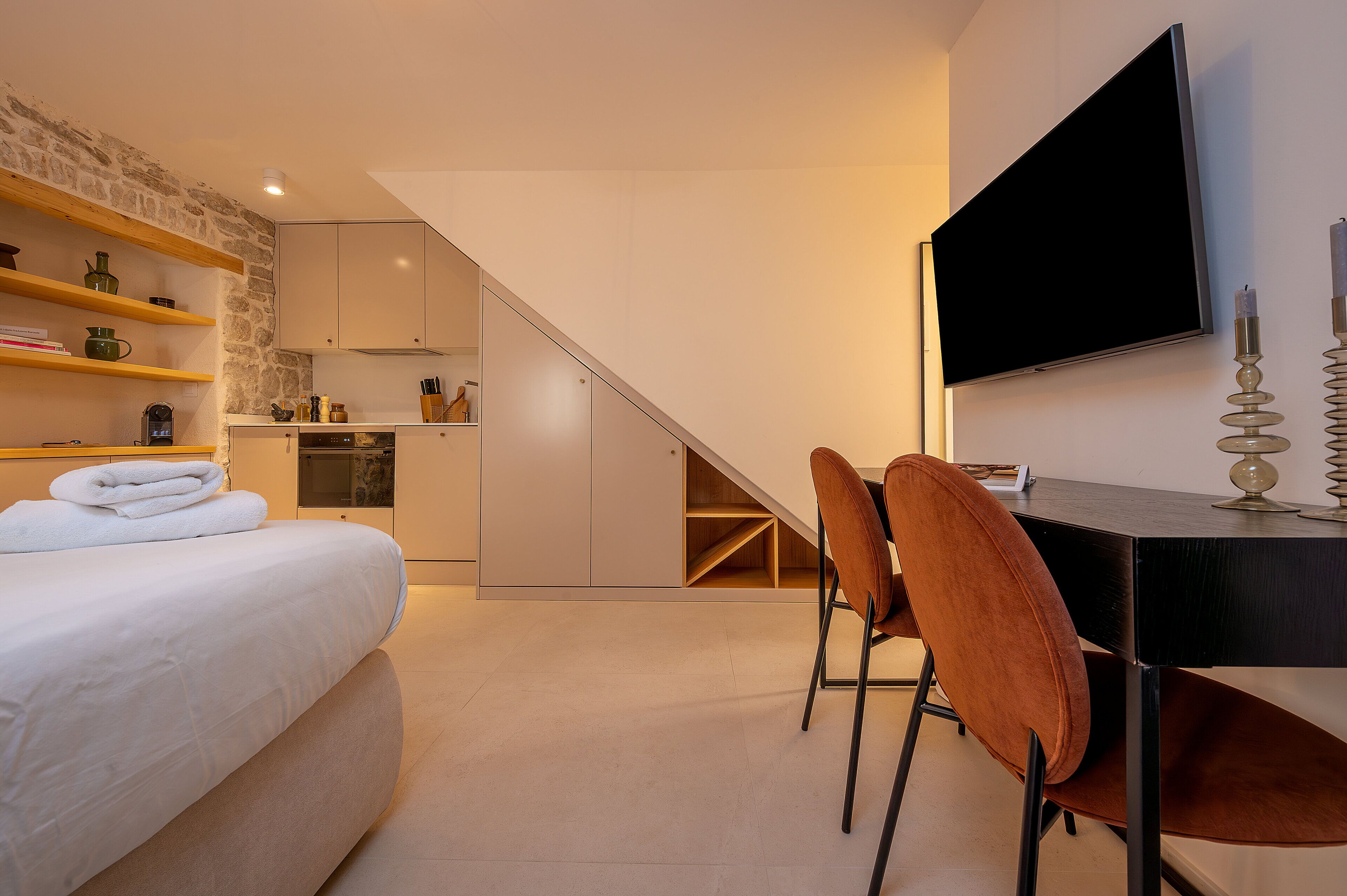 A modern hotel room with a bed, a dining table with chairs, a TV, and a kitchen area.