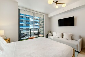 A hotel room with a large bed, a sofa, a TV, and a balcony with a view of a blue building. - Crosby Miami WTC Studio Retreat #408 (Miami)