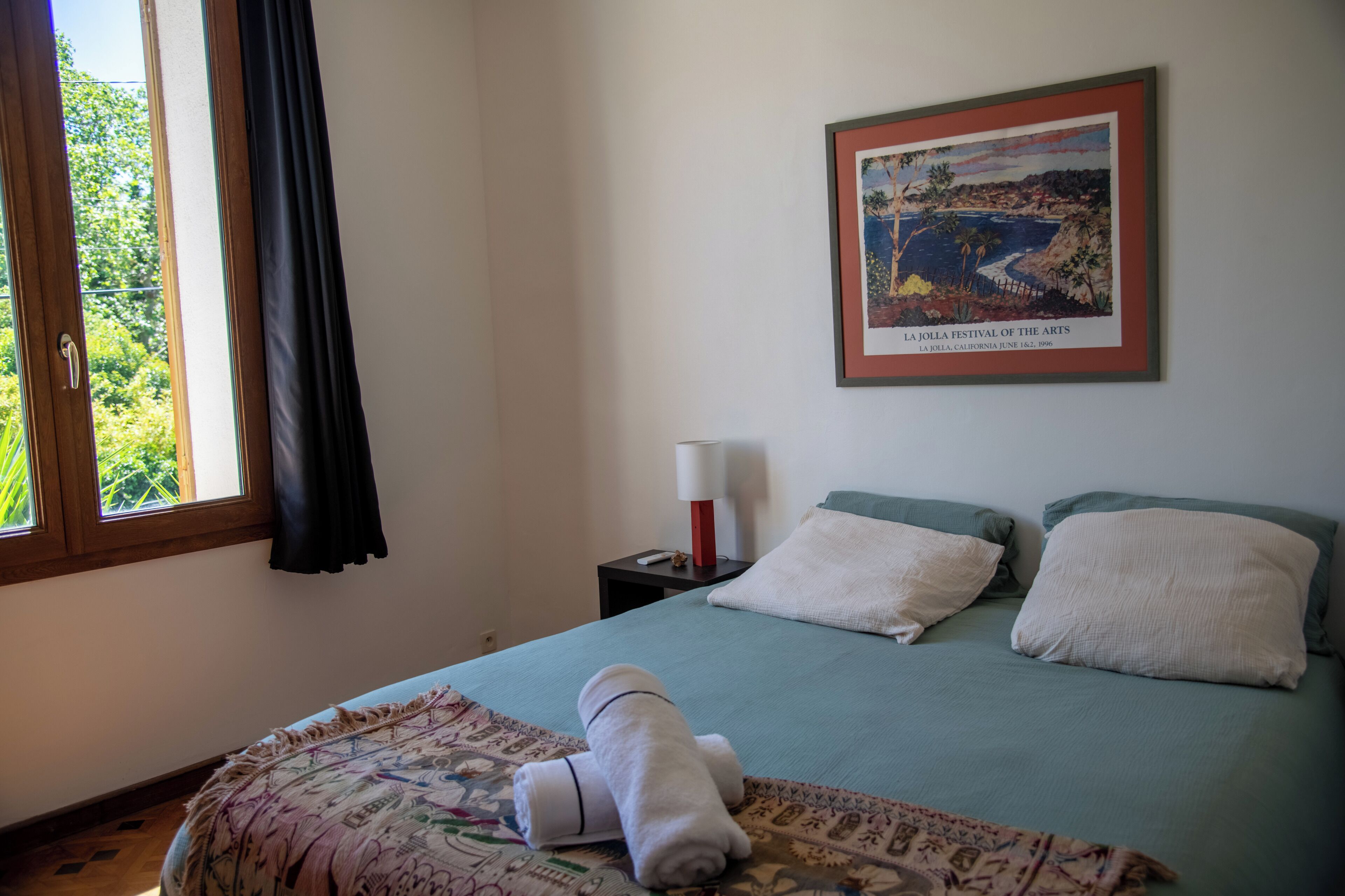A bedroom with a bed, pillows, a nightstand with a lamp, a framed picture on the wall, and a window with curtains.