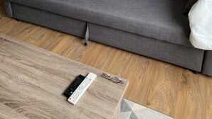 Een grijze bank met kussens, een houten salontafel met een afstandsbediening, en een stoel.