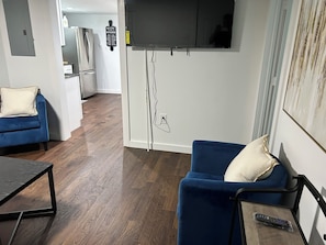 A living room with a TV mounted on the wall, a sofa, a coffee table, and a kitchen area in the background.