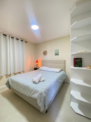 Ein ordentlich bezogenes Bett mit gemustertem Überwurf, eine Nachttischlampe, ein Buch auf einem Regal und eine dekorative Wandplatte.