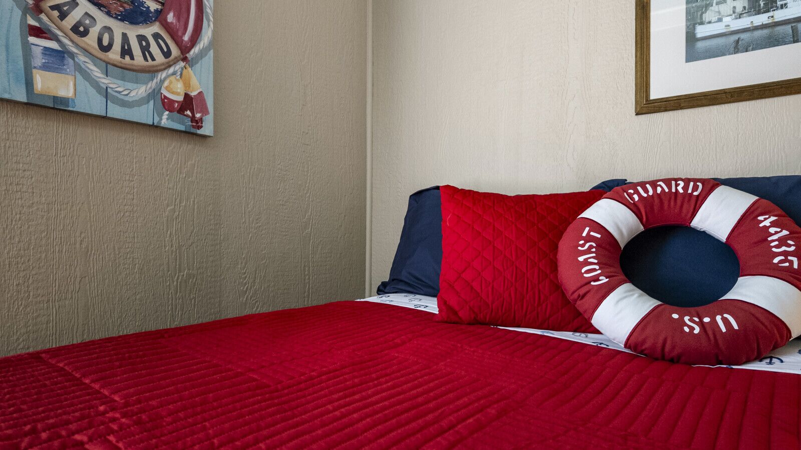 Una cama con un edredón rojo, un almohadón con diseño de boia salvavidas y una pared con un cuadro enmarcado.