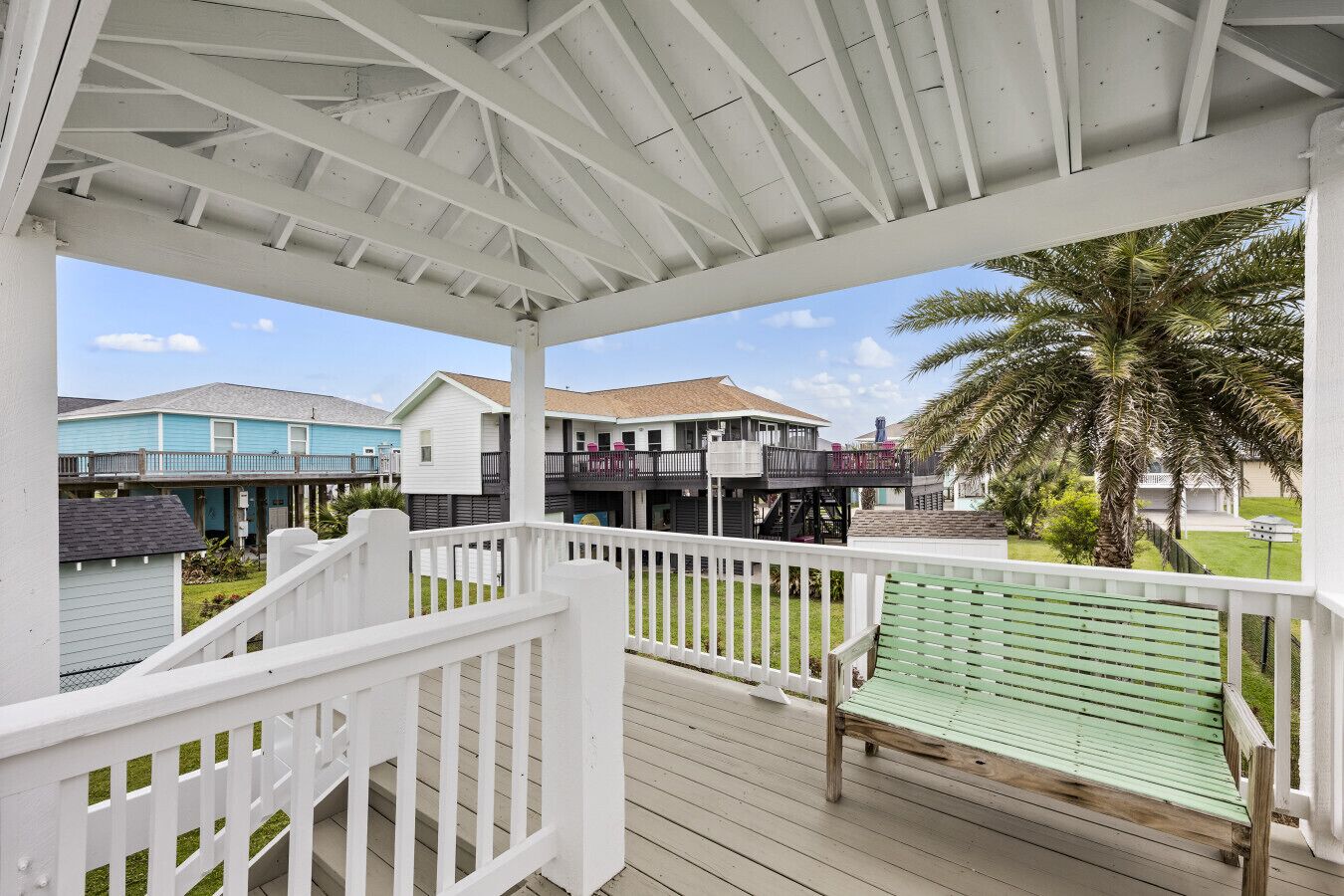 A covered porch with a white railing, a green bench, and a view of houses and palm trees.