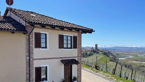 A two-story building with a tiled roof, a balcony with a table and chairs, and a view of a vineyard and mountains in the background.