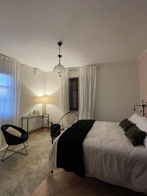 Ein Schlafzimmer mit Bett, Stuhl, Schreibtisch, Lampe und einem Fenster mit Vorhängen.