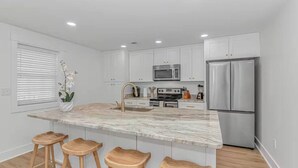 Una cucina moderna con mobili bianchi, un'isola in marmo, elettrodomestici in acciaio inossidabile e sgabelli in legno.