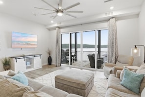 A spacious living room with a large TV, comfortable seating, a ceiling fan, and a balcony with a view.