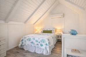 A bedroom with a sloped ceiling, a bed with a patterned bedspread, bedside lamps, a small shelf with decorative items, and an air conditioning unit.