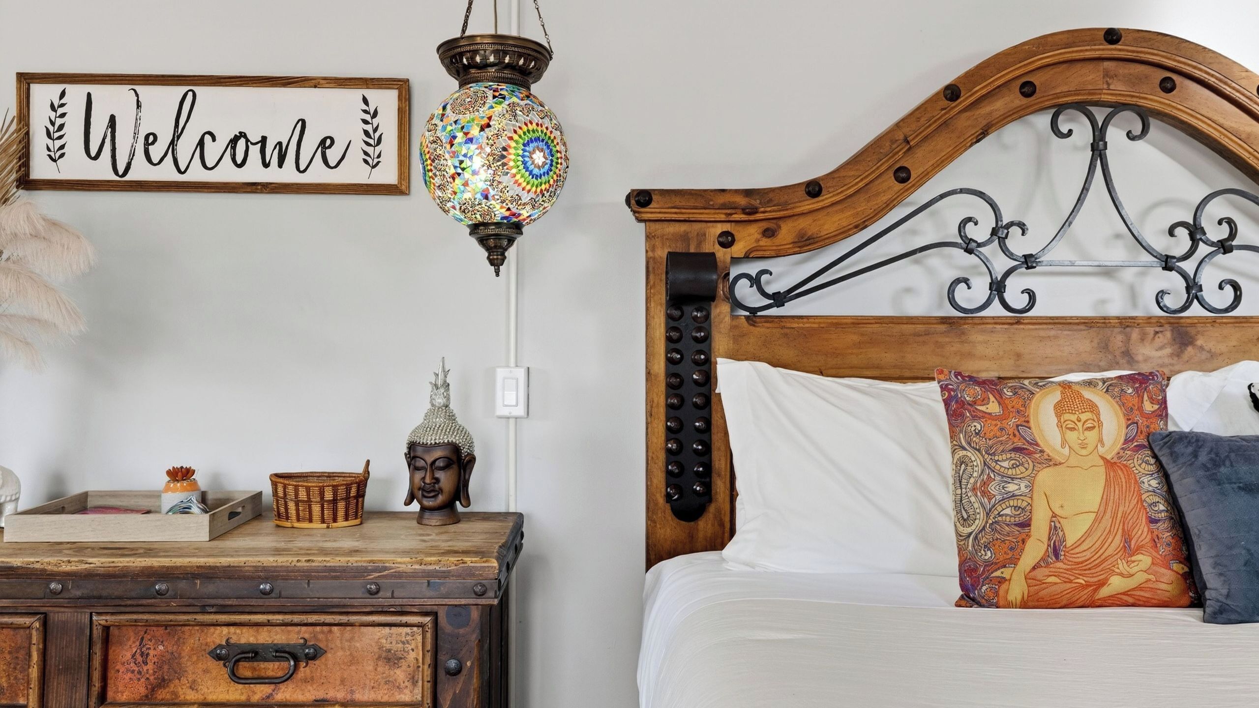 Un dormitorio con cabecera de madera, un letrero de bienvenida, una lámpara decorativa, una estatua de Buda y una mesita de noche con una cesta y una figurita.