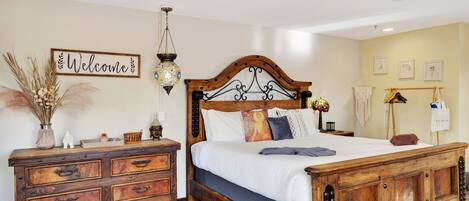 A bedroom with a wooden bed, a dresser, a 'Welcome' sign, a hanging lamp, and a macrame wall hanging.