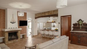A spacious living area with a fireplace, a dining area with chairs, a kitchen with a bar, and a large TV mounted on the wall. - Quinta Alegria - Rustic Cottage (Alcobaça)