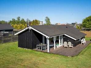 Un cobertizo de metal negro con techo inclinado, una terraza de madera con mobiliario exterior y un cielo despejado.