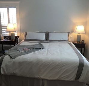 Una cama bien hecha con almohadas, una mesita de noche con una lámpara y una ventana con persianas.