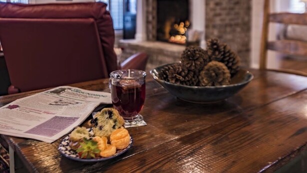 A dining area with a fireplace, a table with a newspaper, a glass of red drink, and a bowl of cookies.